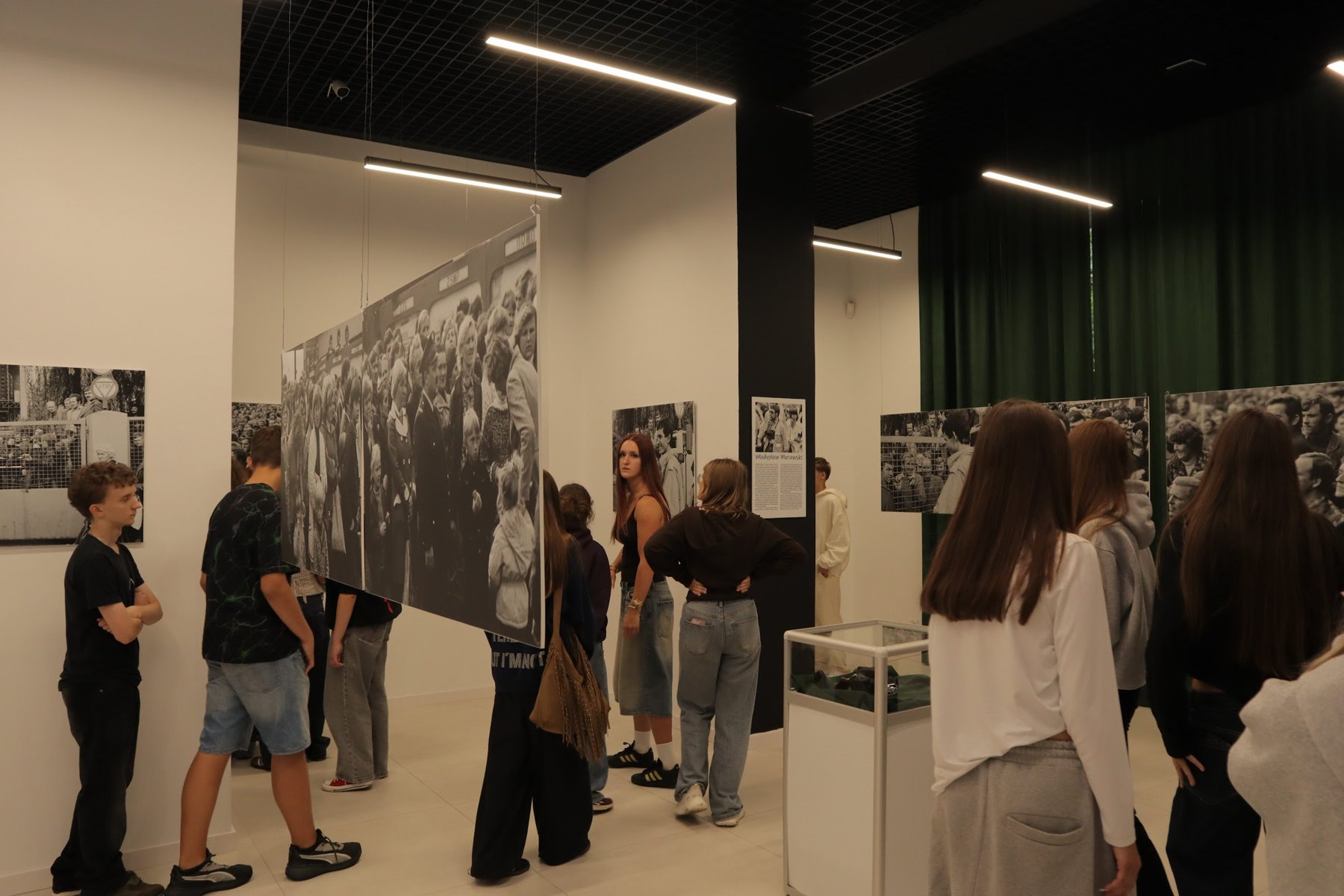 Sala wystawowa z dużymi czarno-białymi fotografiami na białych ścianach. Młodzież ogląda plansze, większość osób stoi tyłem.