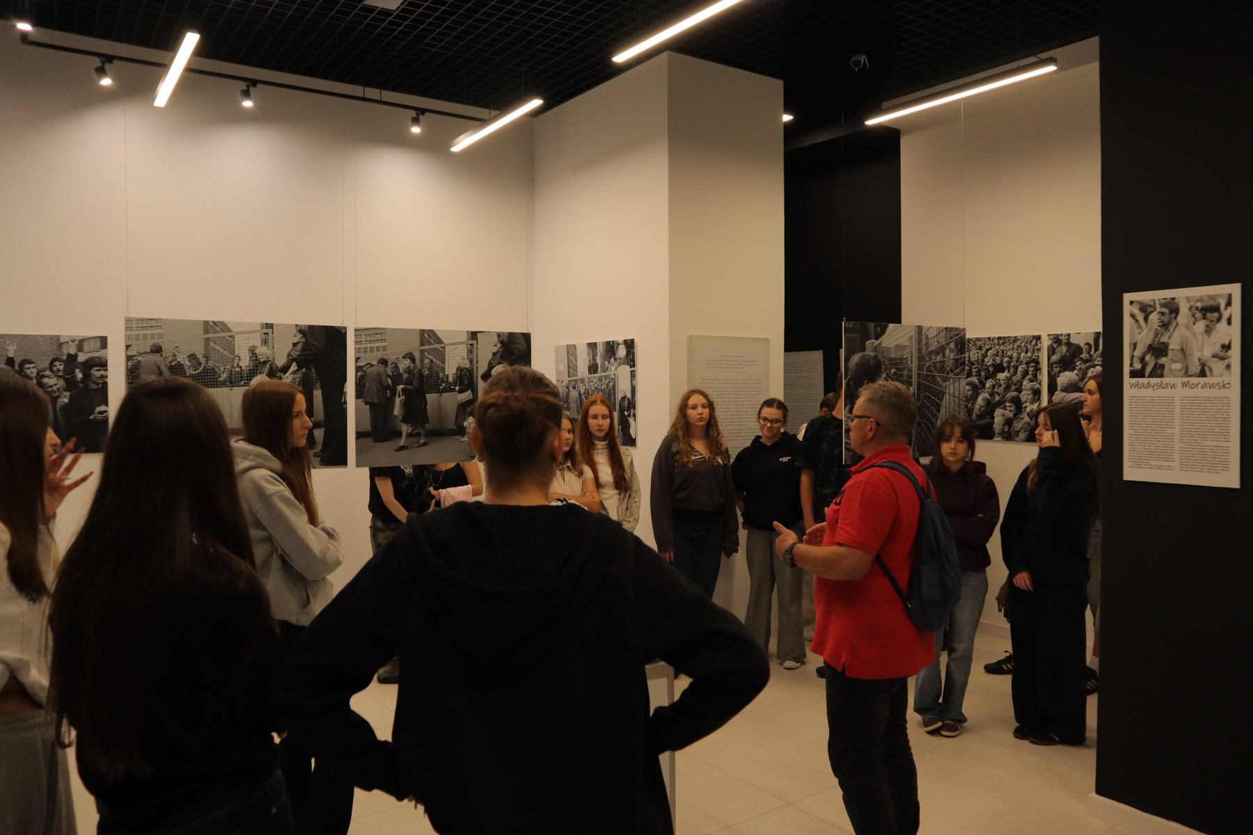 Grupa młodzieży stoi w sali wystawowej wokół przewodnika w czerwonej koszulce. Na ścianach czarno-białe fotografie i plansze z tekstem.