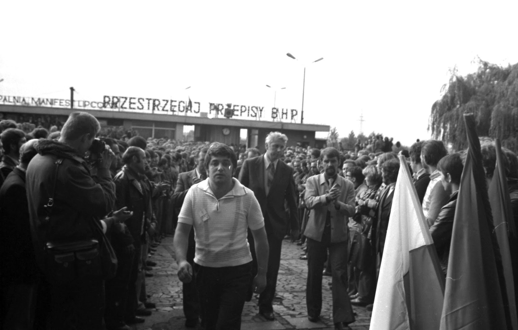Trzej mężczyźni idą środkiem szpaleru utworzonego przez tłum. Po lewej biało-czerwone flagi, nad bramą napis „Przestrzegaj przepisy BHP”.