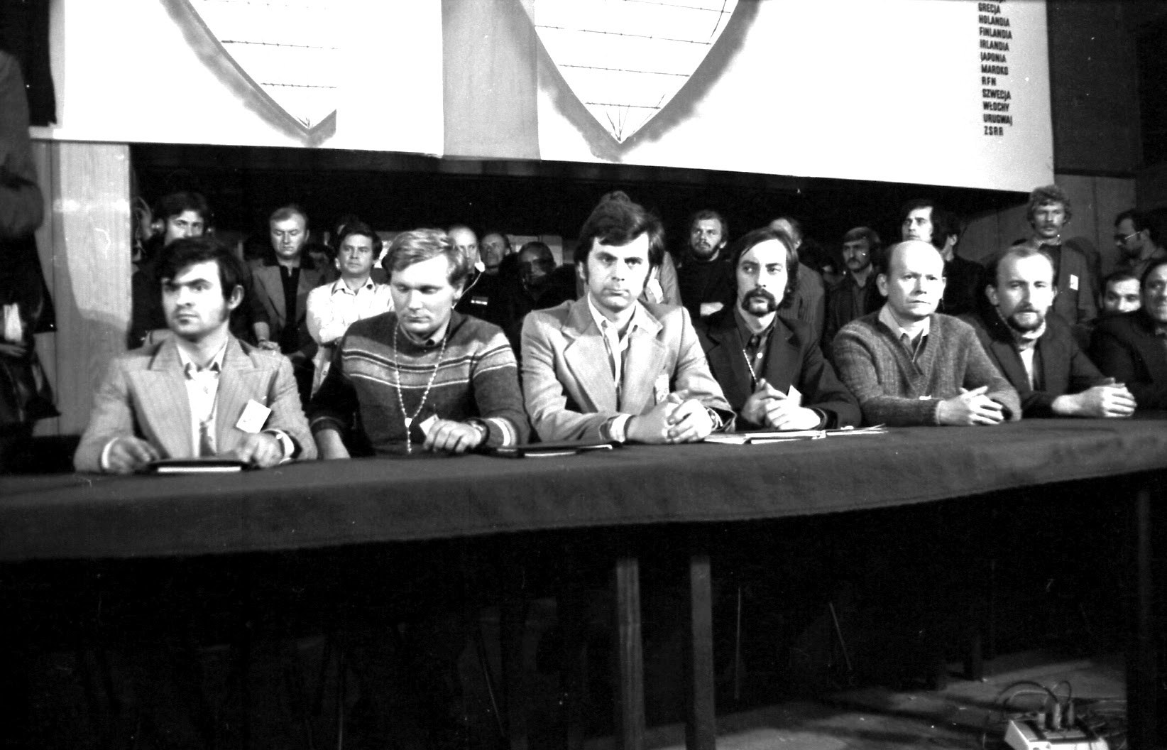 Sześciu młodych mężczyzn z identyfikatorami siedzi za długim stołem nakrytym ciemnym suknem. Za nimi stoją kolejni uczestnicy.