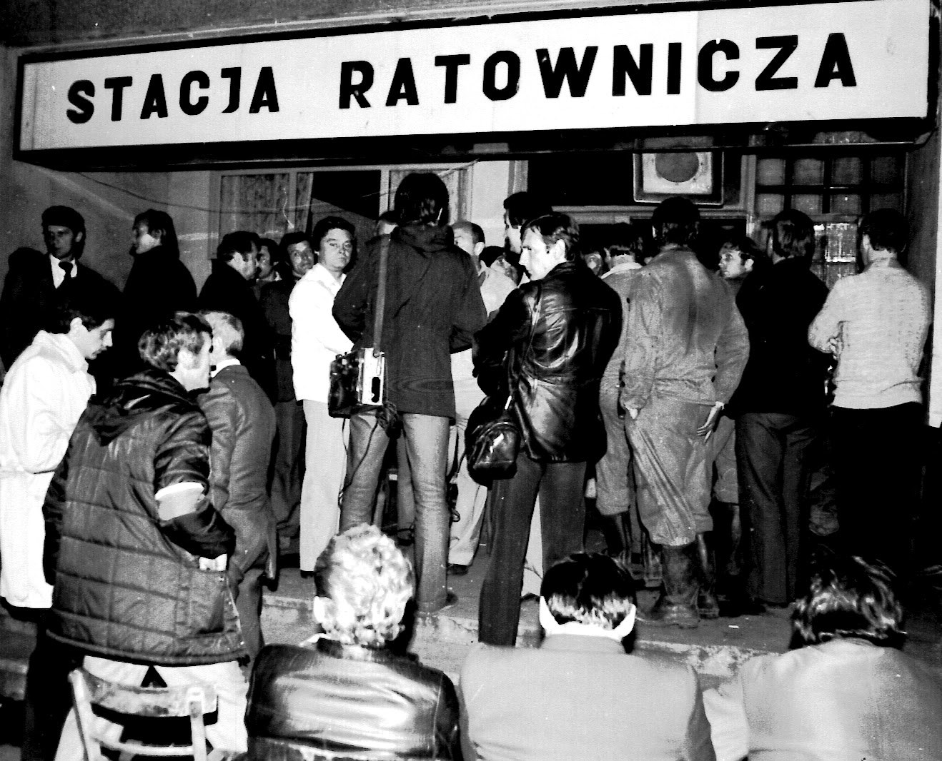 Grupa mężczyzn stoi wieczorem pod dużym napisem „Stacja Ratownicza”. Na pierwszym planie siedzący tyłem ludzie i puste krzesło.