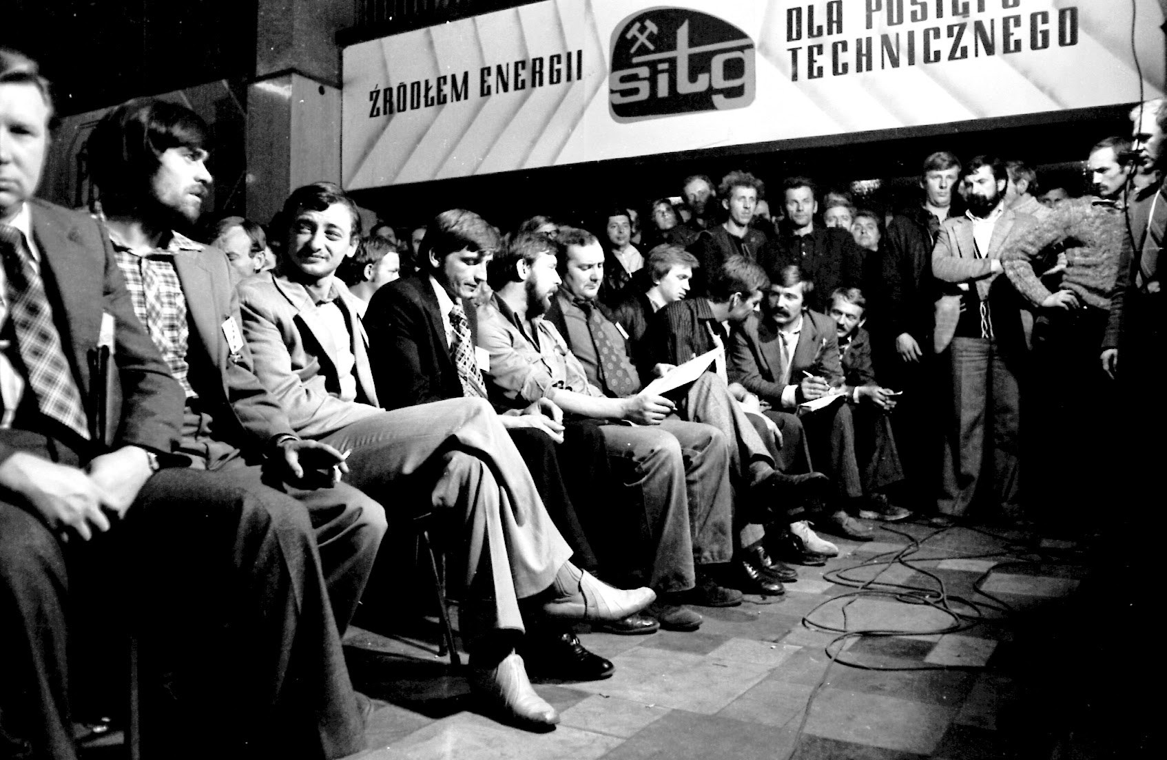 Rząd mężczyzn siedzi na krzesłach, jeden czyta z kartki. Za nimi stoją inni, nad nimi tablica z napisem „…dla postępu technicznego”.