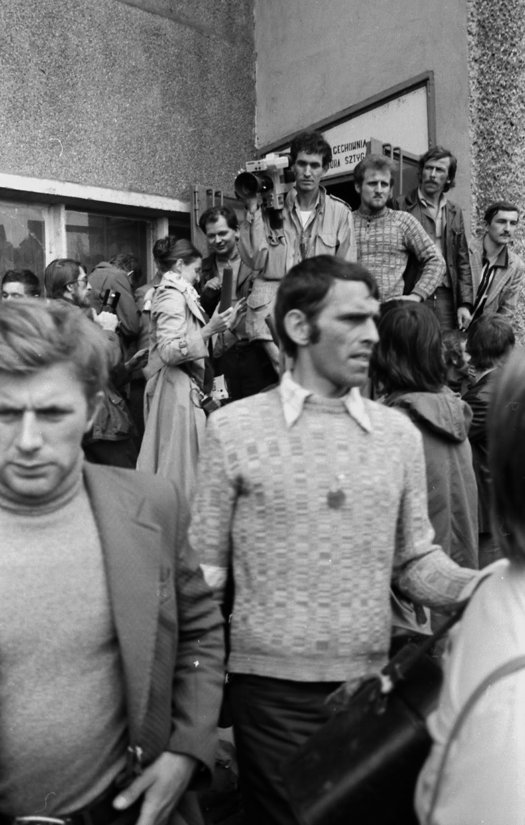 Mężczyzna w swetrze idzie przez tłum. Za nim operator z kamerą filmową i grupa ludzi przy budynku.
