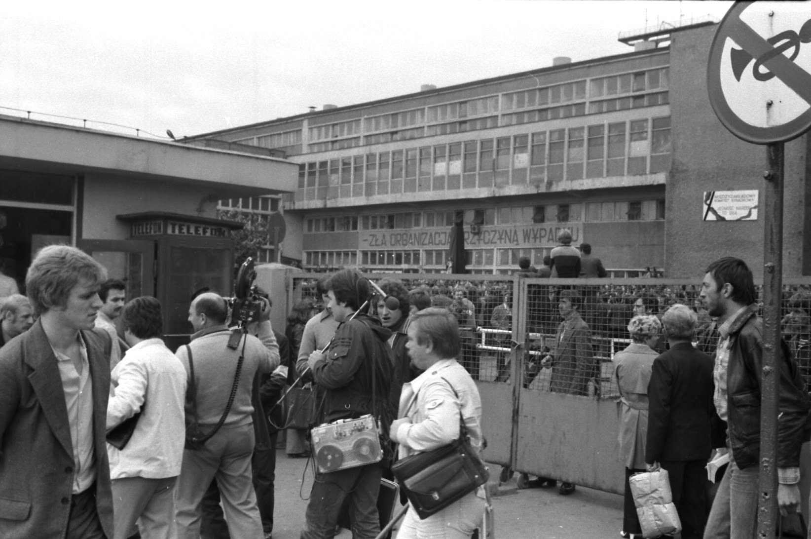 Grupa dziennikarzy z magnetofonem i kamerą stoi przy ogrodzeniu. Za siatką tłum na dziedzińcu i budynek zakładu.