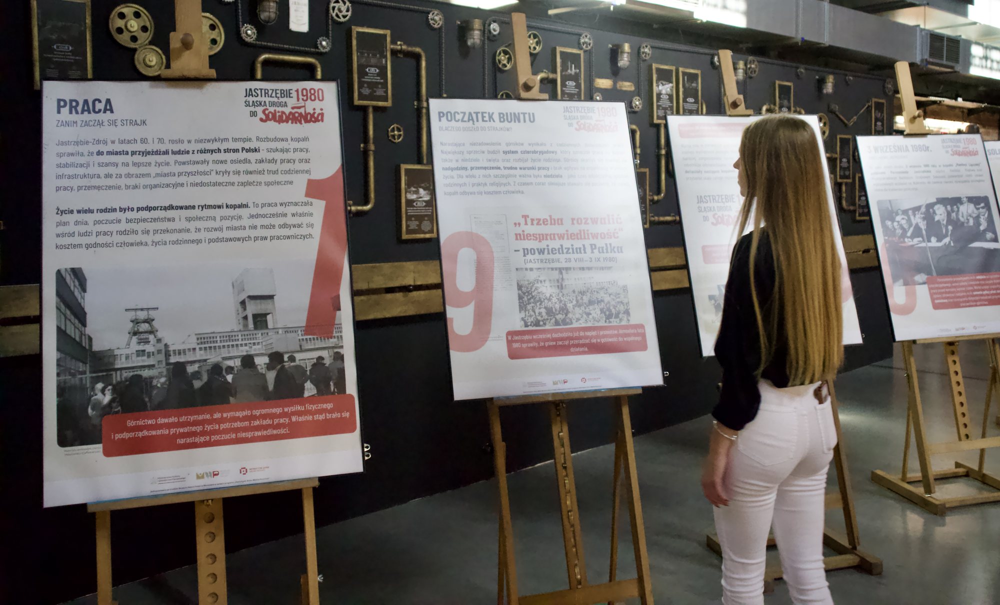 Rząd plansz na drewnianych sztalugach pod ciemną ścianą. Na pierwszej tytuł „Praca” i duża czerwona cyfra 1, na drugiej „Początek buntu” i cyfra 9. Przy trzeciej planszy stoi kobieta i czyta.