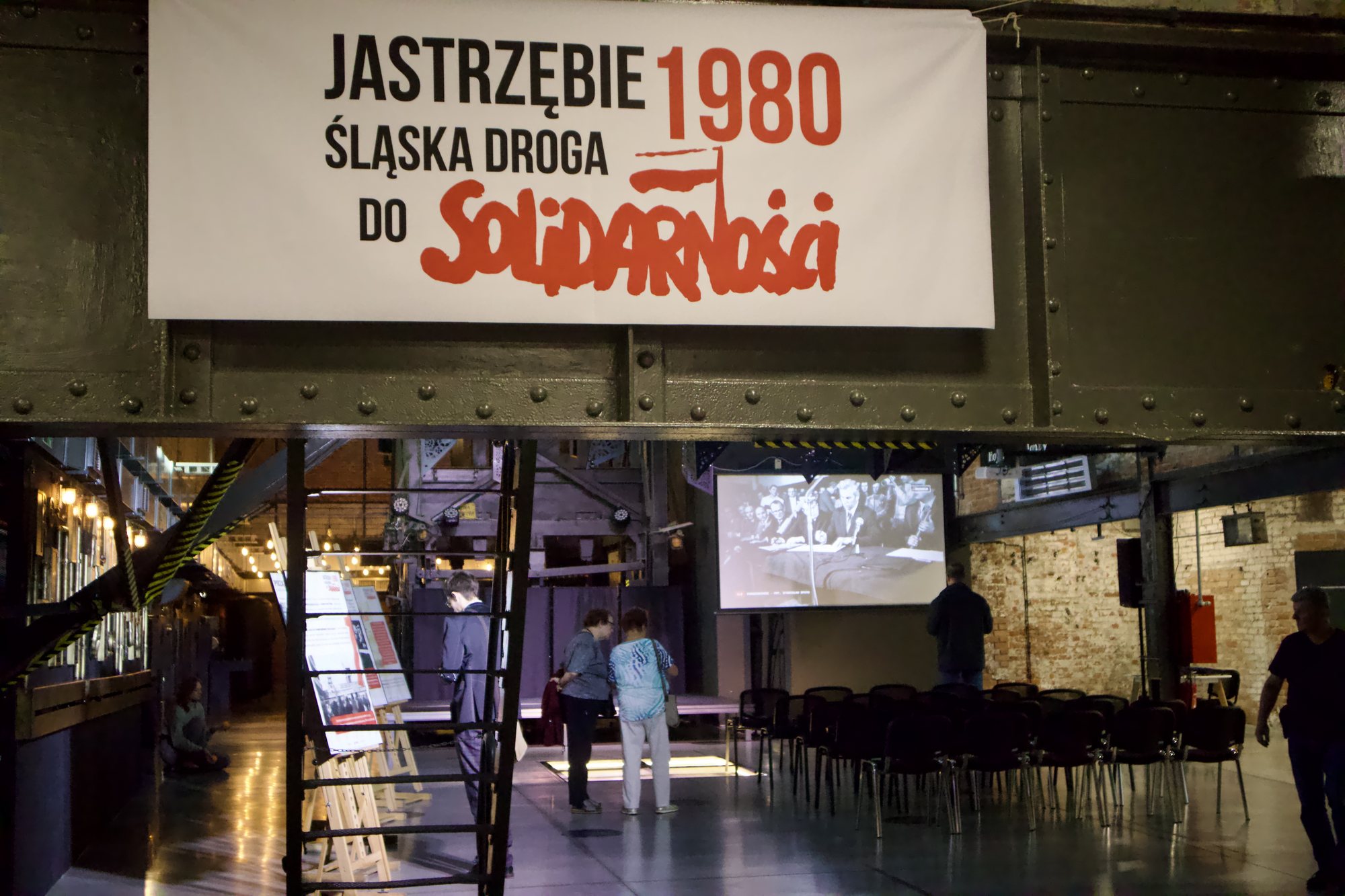 Hala kopalni widziana z antresoli: u góry baner „Jastrzębie 1980. Śląska droga do Solidarności”, niżej stalowe schody, rzędy czarnych krzeseł i ekran z czarno-białą fotografią. Po lewej widać rząd plansz na sztalugach.
