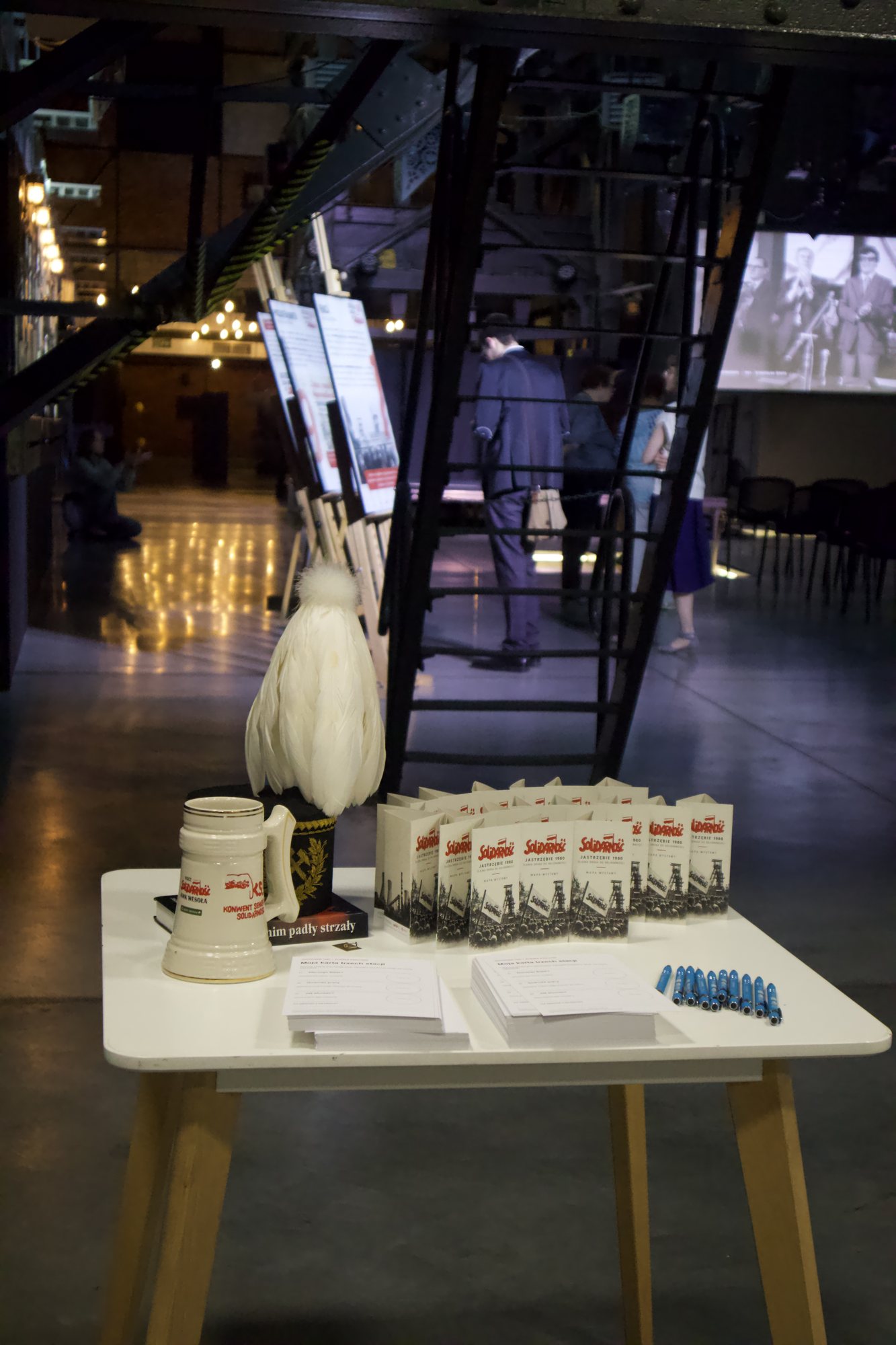 Biały stolik, na nim górnicza czapka galowa z białym pióropuszem, ceramiczny kufel, rząd złożonych folderów z napisem „Solidarność. Jastrzębie 1980”, dwa stosy kart uczestnika i niebieskie długopisy. W tle stalowe schody, plansze na sztalugach i ekran.