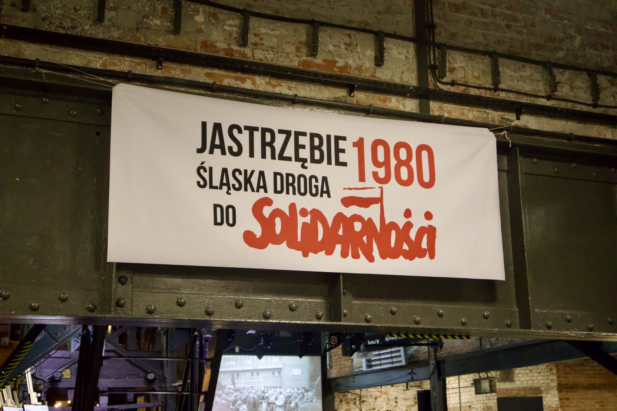 Biały baner z napisem „Jastrzębie 1980. Śląska droga do Solidarności” zawieszony na nitowanej stalowej konstrukcji w hali Zabytkowej Kopalni Ignacy. W tle ceglana ściana.