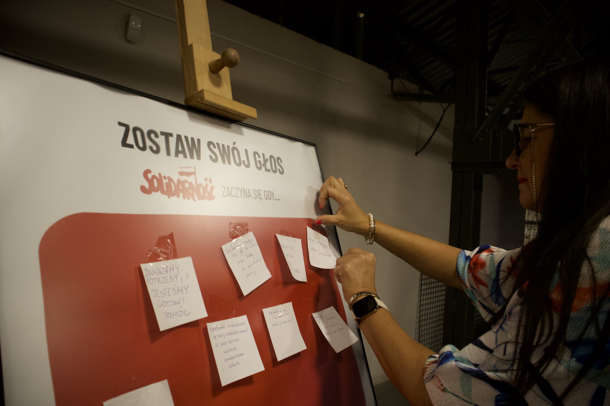 Kobieta przykleja karteczkę do czerwonego dymka na planszy „Zostaw swój głos”. Na dymku wisi już kilka zapisanych karteczek.