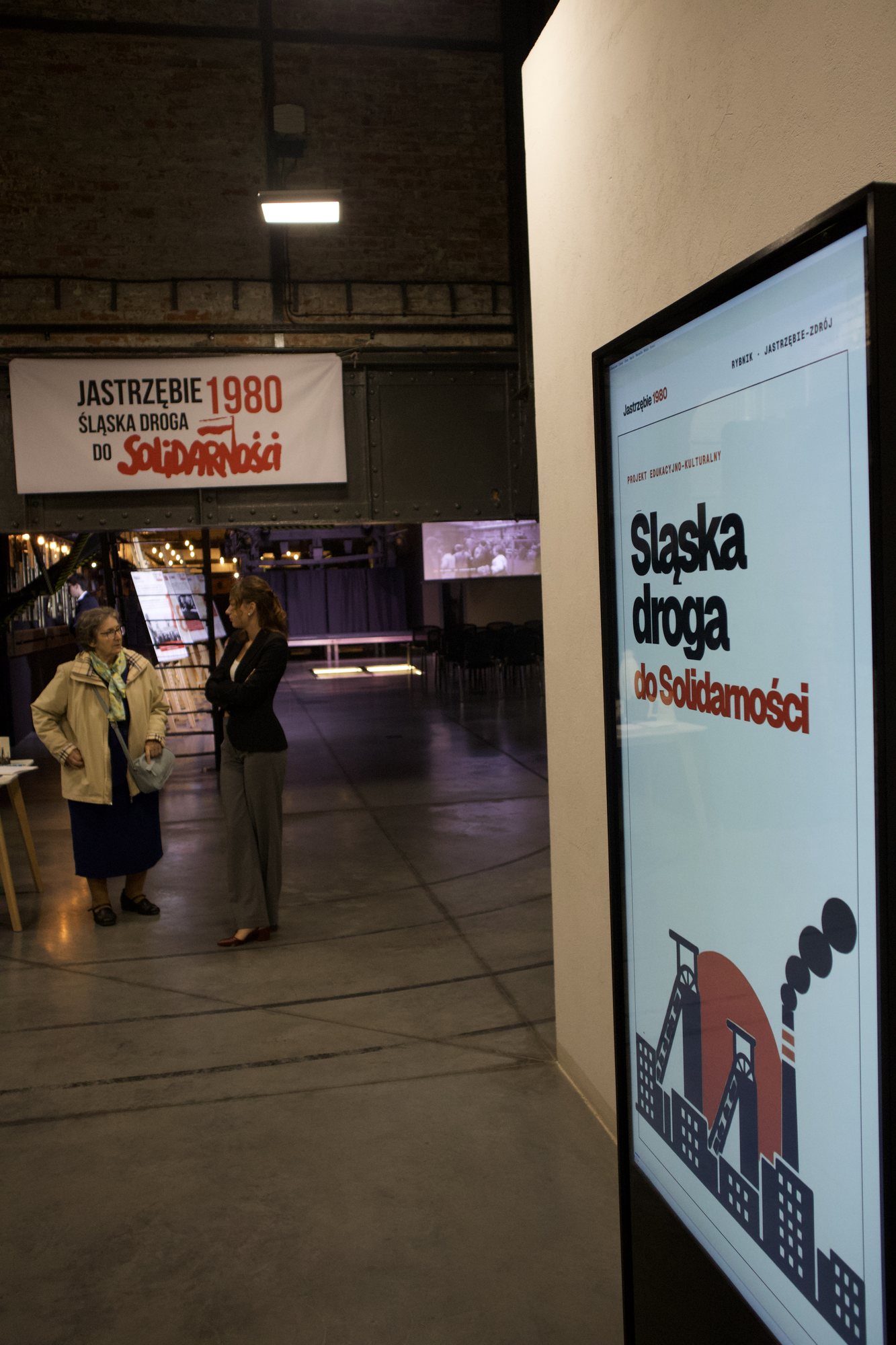 Pionowy ekran z afiszem „Śląska droga do Solidarności”; w głębi hali widać baner projektu i dwie osoby.