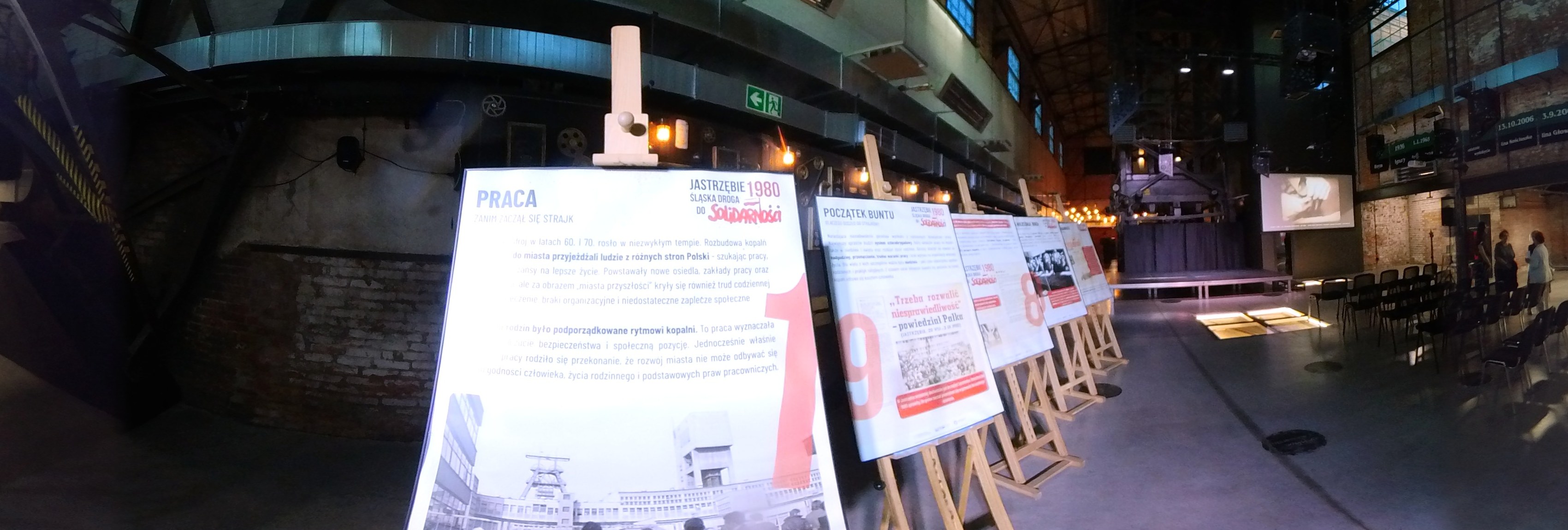 Szeroki wycinek panoramy 360° hali: na pierwszym planie plansza „Praca” na sztaludze, za nią kolejne plansze, w głębi scena, ekran i krzesła widowni.