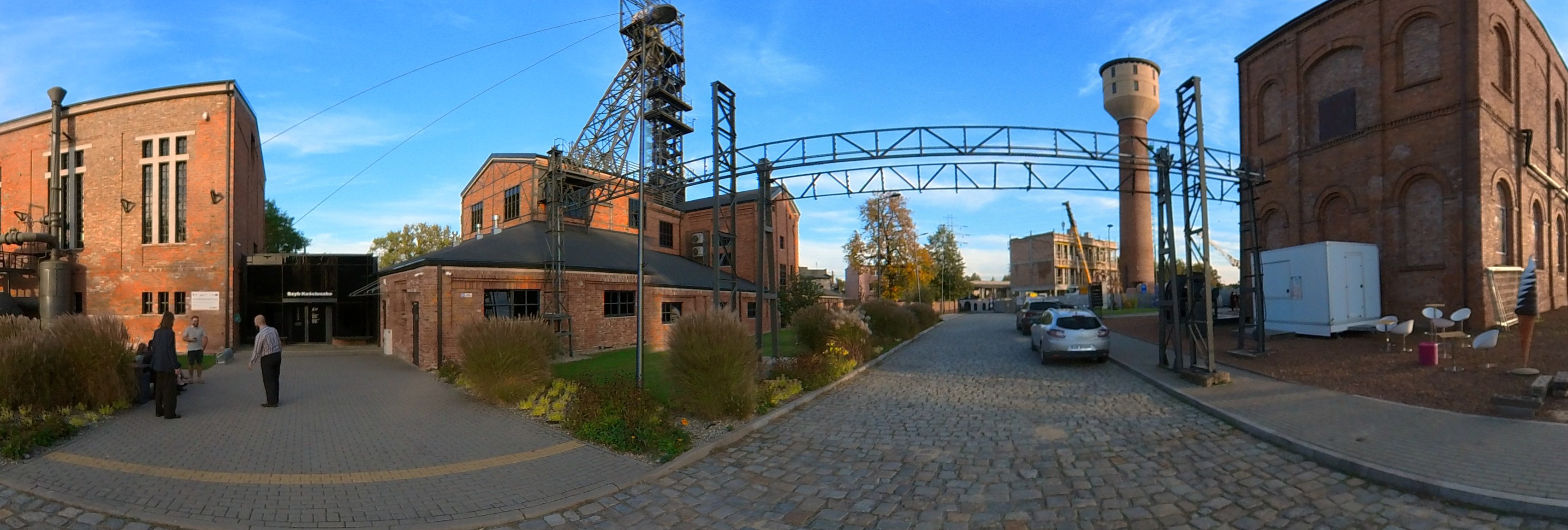 Szeroki wycinek panoramy 360° placu przed wejściem: ceglane budynki kopalni, stalowa brama, wieża szybowa i brukowany plac.