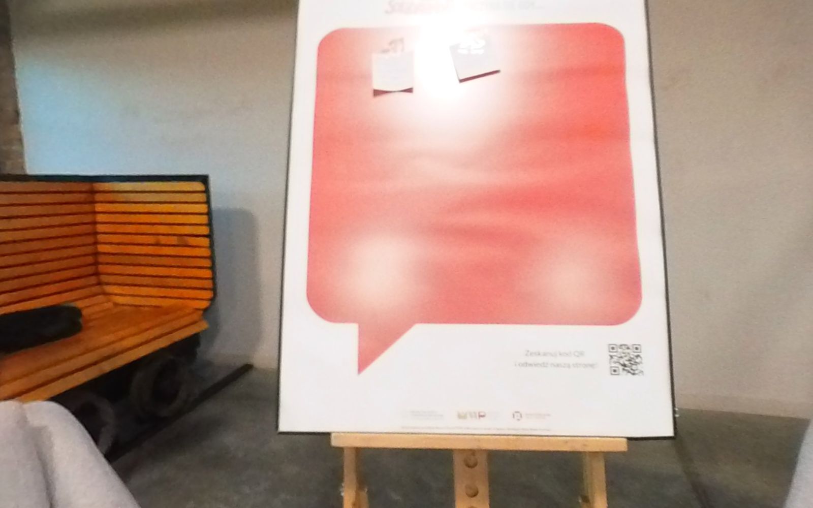 Plansza „Zostaw swój głos” z czerwonym dymkiem na sztaludze, między szarą kanapą a fotelem.