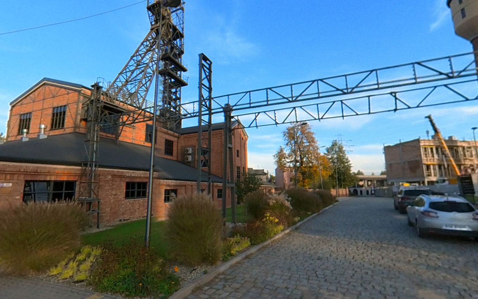 Brukowany plac i droga przy ceglanym budynku kopalni; nad dachem stalowa wieża szybowa, po lewej łukowa brama.