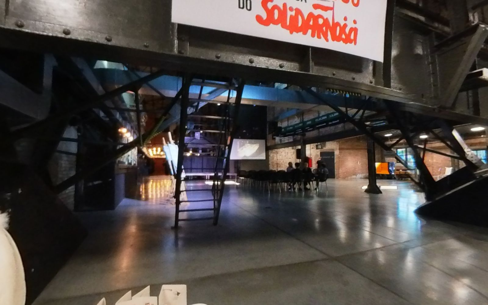 Baner „Jastrzębie 1980. Śląska droga do Solidarności” nad wejściem do hali; na pierwszym planie biały stolik z długopisami i materiałami projektu.